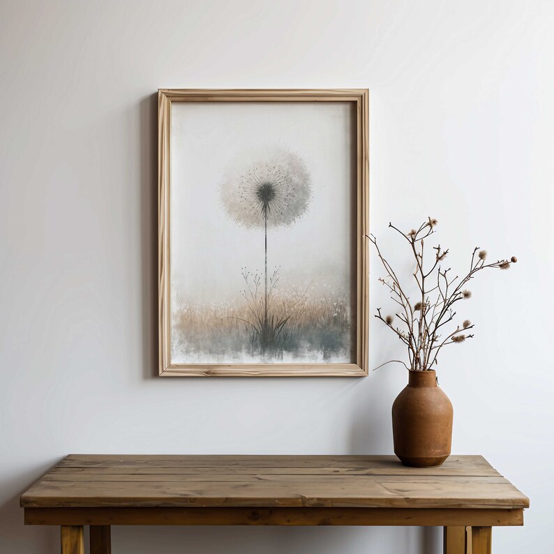 May include: A framed print of a watercolour painting of a dandelion with a soft, muted colour palette. The dandelion is in the centre of the print and is surrounded by a field of grass. The print is in a wooden frame and is hanging on a white wall.