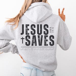 May include: A light gray hoodie with the text "JESUS SAVES" in large black letters on the back, along with the Bible verse John 14:6. The hoodie has a drawstring hood and a kangaroo pocket.