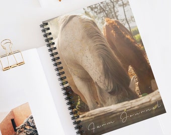 May include: A white and brown horse with a long mane and tail is grazing in a field. The image is used as the cover of a spiral bound journal with the text "Farm Journal" written in cursive on the bottom right corner.