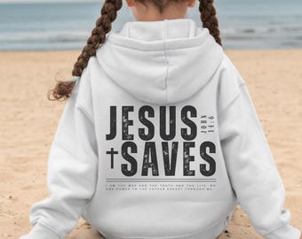 May include: White hooded sweatshirt with the text "JESUS + SAVES" in black on the back. The sweatshirt also features the Bible verse John 14:6. The wearer is sitting on a sandy beach.