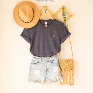 May include: A navy blue t-shirt with the text "FINDING GROWTH" and a leaf design, paired with denim shorts, a tan hat, and a woven crossbody bag. The shirt is hanging on a wooden hanger.
