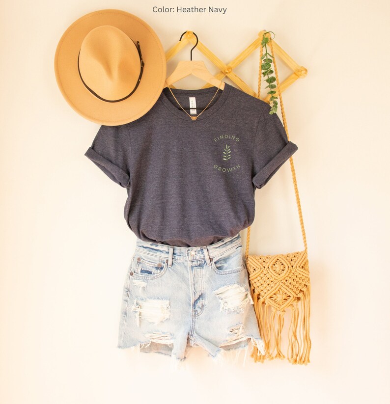 May include: A navy blue t-shirt with the text "FINDING GROWTH" and a leaf design, paired with denim shorts, a tan hat, and a woven crossbody bag. The shirt is hanging on a wooden hanger.
