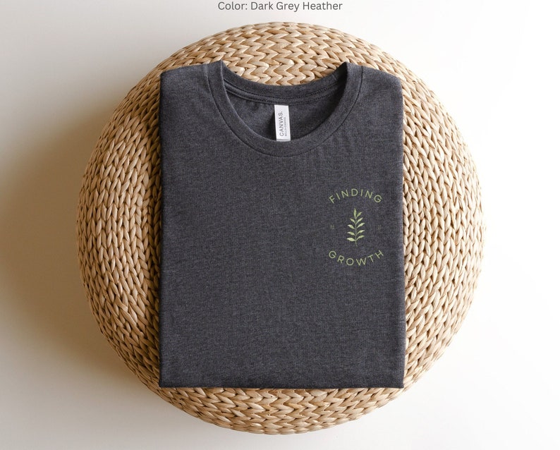 May include: Dark grey heather t-shirt with a crew neck, folded neatly on a woven surface. The shirt features the text "FINDING GROWTH" in a circular design with a small leaf illustration. The shirt is made of a soft material.