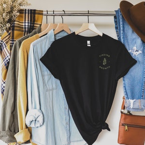 May include: A black t-shirt with the words "Finding Growth" and a small leaf design printed in green. The shirt hangs on a clothes rail with other shirts and a pair of blue jeans. A brown hat and purse are also visible.