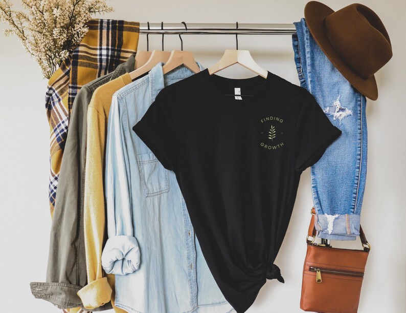 May include: A black t-shirt with the words "Finding Growth" and a small leaf design printed in green. The shirt hangs on a clothes rail with other shirts and a pair of blue jeans. A brown hat and purse are also visible.