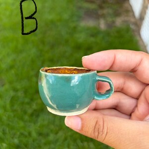 May include: A miniature ceramic cup with a glossy teal exterior and a textured, golden-brown interior. The cup features a small handle on the right side and a white base. The letter B is written in black ink in the upper left corner of the image.