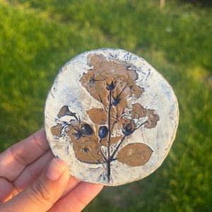 May include: A circular, flat clay disc featuring a pressed botanical design. The center displays a brown, textured imprint of a plant with leaves and small berries, while the surrounding border is finished in a distressed, textured white glaze.
