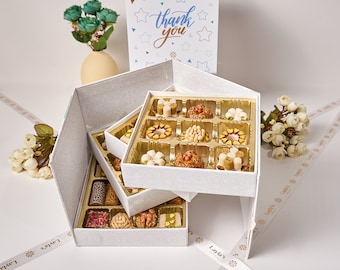 May include: A white gift box with a layered design, containing a variety of small, sweet treats. The box is decorated with a delicate pattern and the top layer features a card with the words "thank you" in blue lettering.