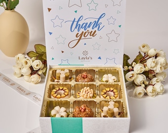 May include: A white gift box with a blue and white star pattern and the words "thank you" on the lid. The box contains nine small, round, decorated treats in a gold tray. The box is surrounded by white flowers.