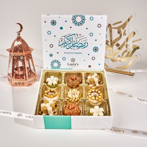 May include: A white box with a blue and white design and the text "Ramadan Kareem" and "Layla's Delicacies". The box contains nine small, round, colorful pastries. The box is sitting on a white surface with a gold decorative lantern and a gold decorative sign.