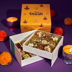May include: A festive gift box for Diwali, featuring an open box of assorted sweets. The box is decorated with a gold and white pattern and the words "Happy Diwali Festival of Lights." Candles and orange pom-poms surround the box.