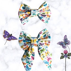 May include: Two butterfly-patterned fabric bow ties. The smaller bow tie is a traditional bow tie shape. The larger bow tie is a more modern style with long tails. Both bow ties are white with a colorful butterfly print.