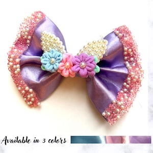 May include: A lavender satin hair bow with a beaded edge and a cluster of small fabric flowers in blue, pink, and purple. The bow is decorated with faux pearls and the text "Available in 3 colors".