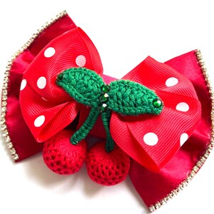 May include: A red hair bow with white polka dots, featuring a central embellishment of two crocheted red cherries with green leaves and stems. The edges of the bow are lined with a row of small, clear, sparkling rhinestones.