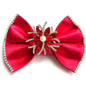 Könnte beinhalten: Eine leuchtend rote Satinschleife mit glitzerndem Strassbesatz. Die Schleife hat eine zentrale florale Verzierung mit roten Edelsteinen und einer Perle in der Mitte, wodurch ein glamouröses Accessoire entsteht.