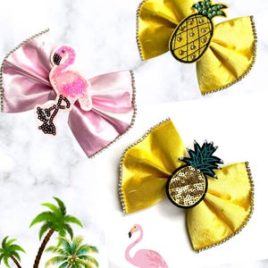 May include: Three hair bows with rhinestone trim. One is light pink with a beaded flamingo patch. Two are yellow, one featuring a beaded pineapple patch with green leaves, and the other a sequined pineapple patch with a dark green leafy top.
