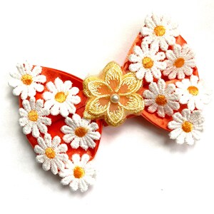 May include: An orange hair bow decorated with white daisy appliqués and a central gold flower with a pearl. This decorative accessory adds a floral touch to any hairstyle. The bow is a charming hair accessory.