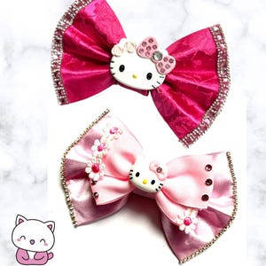 Peut inclure: Deux nœuds pour cheveux avec des centres représentant Hello Kitty. Le nœud supérieur est fuchsia vif avec un motif texturé et des bords en strass. Le nœud inférieur est rose clair avec des petites fleurs blanches, des bords en strass et des strass roses.