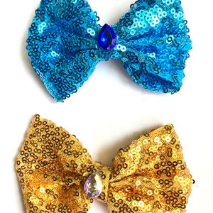Può includere: Due accessori per capelli a fiocco con paillettes. Uno è blu con una gemma blu, l'altro è dorato con una gemma trasparente. I fiocchi sono fatti di piccole paillettes lucenti e sono disposti a forma di fiocco. Le gemme sono a forma di goccia.