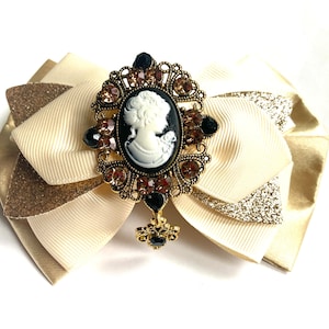 May include: A gold-tone brooch featuring a cameo of a woman's profile in white against a black background. The brooch is surrounded by rhinestones and is attached to a cream-coloured bow with gold glitter accents.