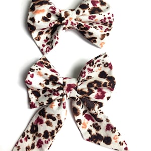 Könnte beinhalten: Zwei Haarbänder mit Leopardenmuster. Das kleinere Band ist ein einfaches Band, während das größere Band zwei lange Schleifen hat.