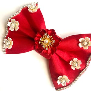 May include: A red satin bow with a large gold and rhinestone flower in the center. The bow is decorated with smaller white pearl flowers and rhinestones around the edges.