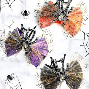 May include: Three hair bows featuring a black glittery spider with a central clear rhinestone, layered over sheer fabric in orange, purple, and grey. Each bow is accented with gold-colored spider web netting and small rhinestones.
