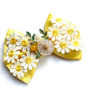 May include: A yellow satin bow with white daisy lace trim and a floral brooch with a clear crystal centre and yellow and green accents.