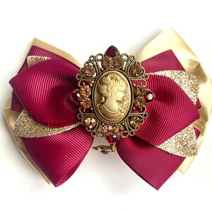 May include: A burgundy and gold bow with a cameo brooch featuring a woman's profile. The brooch is adorned with rhinestones and has a gold finish.