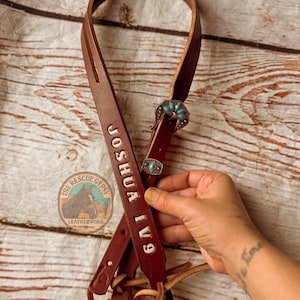 Puede incluir: Una brida de cuero marrón con el nombre "JOSHUA IV9" grabado en blanco. La brida presenta una hebilla decorativa plateada con detalles turquesas. El logotipo de The Rescue Gypsy Leatherwork es visible.