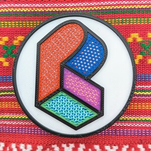 May include: A white circular pin with a black border and a multi-colored geometric design. The design features a large, stylized letter "R" with a red, blue, purple, and green pattern.