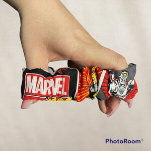 May include: A Marvel-themed scrunchie featuring the Marvel logo and comic book character designs. The scrunchie is made of fabric with a black, red, yellow, and gray color scheme. The scrunchie is designed to hold hair in place.