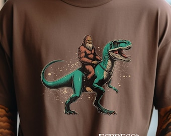 Può includere: Una maglietta marrone con una stampa grafica di un Bigfoot che cavalca un dinosauro verde. Il testo "ESPRESSO" è stampato sul fondo della maglietta.