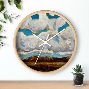 May include: A round wall clock with a light wood frame. The clock face features a landscape painting of a blue sky with fluffy white clouds, brown mountains, and a field. The clock hands are white.