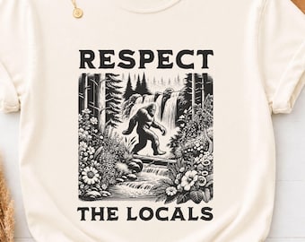 May include: An ivory short-sleeve t-shirt featuring a black graphic print of a forest scene with a waterfall, flowers, and a Bigfoot figure. The text "RESPECT THE LOCALS" is printed in black above and below the central illustration.