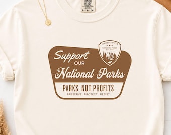 Könnte beinhalten: Ein elfenbeinfarbenes T-Shirt mit einer braunen Grafik, die den Text Support OUR National Parks, ein schildförmiges Emblem mit Berglandschaft sowie die Worte PARKS NOT PROFITS, PRESERVE PROTECT RESIST zeigt. Eine goldfarbene Kette liegt am Ausschnitt.