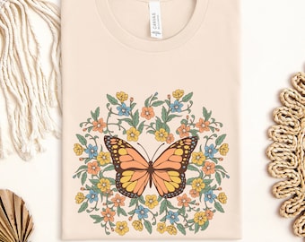 Peut inclure: Un t-shirt crème avec un motif de papillon. Le papillon est orange, jaune et noir, entouré d'une couronne florale avec des fleurs bleues, jaunes et oranges. Le t-shirt a un col rond et le mot "SOFT CREAM" est en bas.