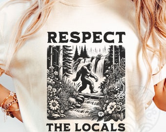 May include: A white t-shirt with a black graphic of a Bigfoot walking through a forest with a waterfall. The text "Respect the Locals" is printed below the graphic.