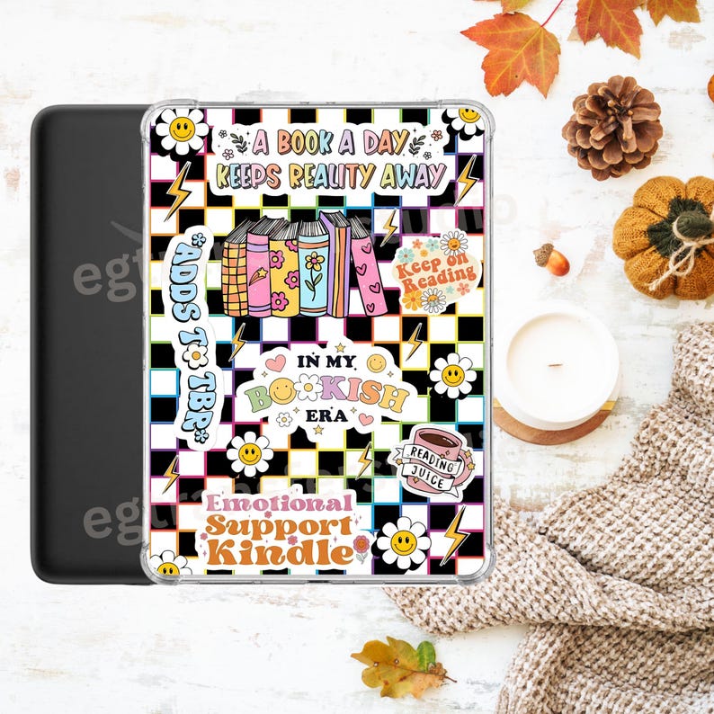 Op de afbeelding: Een zwarte Kindle-hoes met een doorzichtige hoes met een kleurrijk ontwerp. Het ontwerp omvat een geruite achtergrond, boeken, bloemen, bliksemschichten en tekst met de tekst "A Book a Day Keeps Reality Away" en "Emotional Support Kindle."