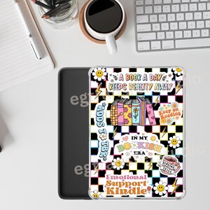 Op de afbeelding: Een doorzichtige Kindle-hoes met een kleurrijk retro-ontwerp. De hoes heeft een zwart-witte geblokte achtergrond met bloemen- en bliksemgrafieken. Tekst bevat "A Book a Day Keeps Reality Away" en "Emotional Support Kindle."