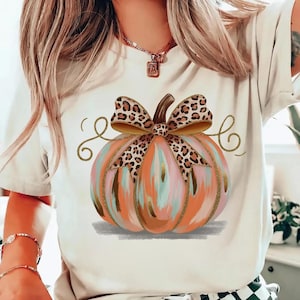 May include: Cream-colored t-shirt featuring a colorful pumpkin design. The pumpkin has orange, pink, and blue brushstrokes, a brown stem, and a leopard print bow. Gold glitter accents the design, perfect for fall.