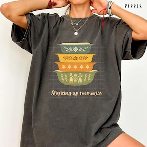 Może przedstawiać: Ciemnoszara koszulka z nadrukiem przedstawiającym stos czterech kolorowych naczyń do zapiekania, każdy z kwiatowymi i ludowymi wzorami. Pod naczyniami wydrukowano napis "Stacking up memories". Słowo "PEPPER" jest wydrukowane w prawym górnym rogu.