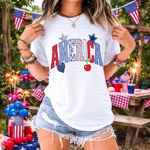 May include: White t-shirt with the word "AMERICA" in red, blue, and patterned fabric letters. The design includes a blue heart, a red apple, and star accents, celebrating patriotic themes. The shirt is paired with denim shorts.