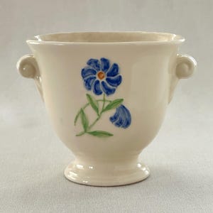 Könnte beinhalten: Ein weißer Keramik-Pflanztopf mit zwei Griffen und einem blauen Blumendesign. Die Blume ist eine blaue Margerite mit gelber Mitte und grünen Blättern.