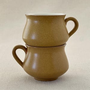 May include: Two brown ceramic mugs stacked on top of each other. The mugs have a rounded shape and a wide handle.