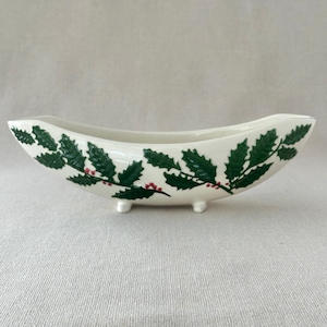 May include: A white ceramic dish with a curved, boat-like shape, resting on three small feet. The dish is decorated with green holly leaves and red berries, creating a festive holiday design. The dish is approximately 10 inches long.