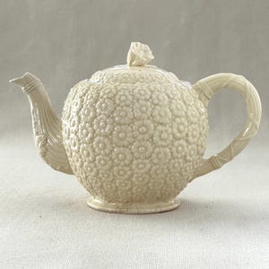 May include: Cream-colored ceramic teapot with a floral pattern. The teapot features a textured surface, a curved handle, and a spout with a decorative design. The lid is topped with a flower-shaped knob. A classic piece for tea lovers.