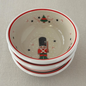 May include: A stack of three ceramic bowls with a festive Christmas theme. Each bowl features a cream-colored interior with a red rim, a soldier graphic, a Christmas tree, and star designs. The bowls are stacked on top of each other.