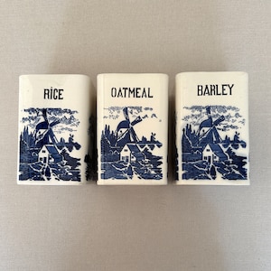 May include: Three white ceramic canisters with blue and white windmill designs. The canisters are labeled "RICE", "OATMEAL", and "BARLEY".