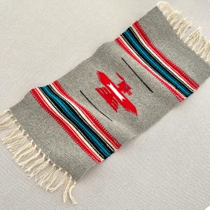 May include: A rectangular gray woven rug with a red bird design in the center. The rug has red, white, and blue stripes on each end, with fringe on the short sides. The rug is approximately 24 inches long and 12 inches wide.